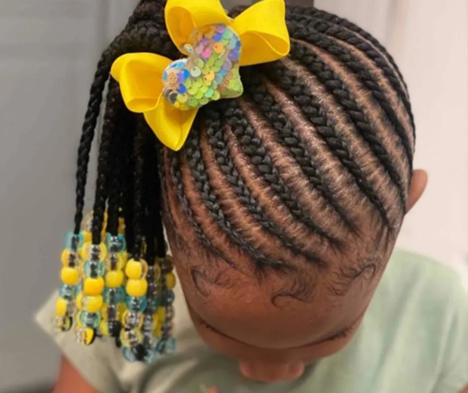A young girl with cornrows styled into a side ponytail adorned with a yellow bow and colorful beads.