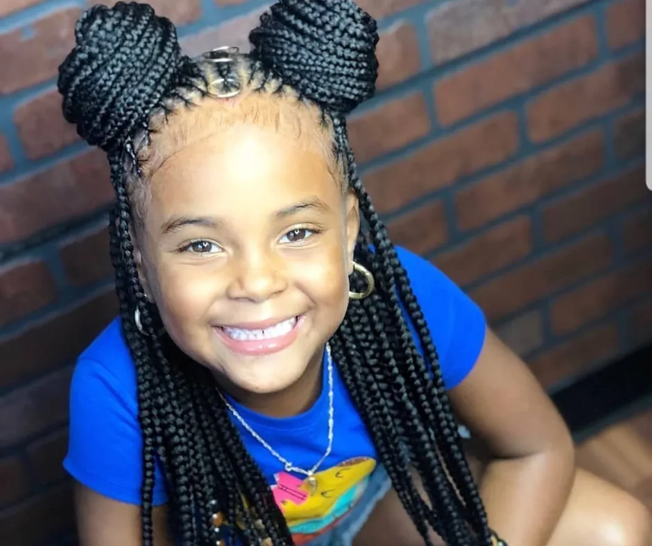 Smiling young girl with knotless braids styled into two high buns and loose strands, wearing a blue shirt.