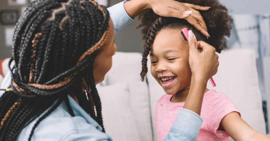 A mother styles her daughter's hair, who is smiling and holding a pink comb, on a couch with light cushions.