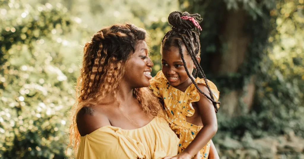 A woman and a young girl with braided hairstyles smile at each other outdoors, both wearing yellow outfits.