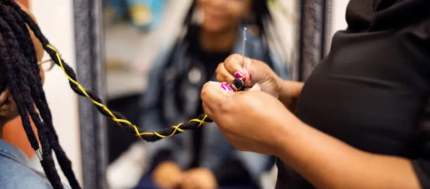 Stylist braiding long faux locs with yellow thread in a hair braiding salon, client in the background.