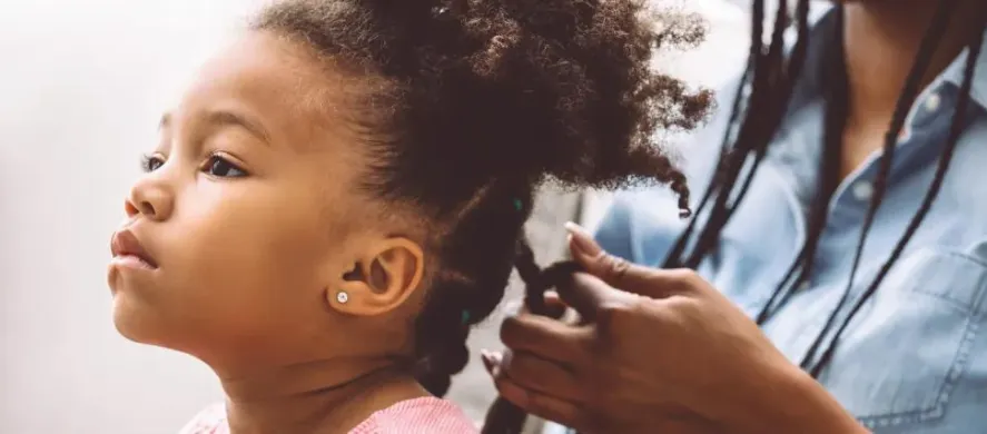 A young girl getting her hair braided by a stylist, wearing a pink top.