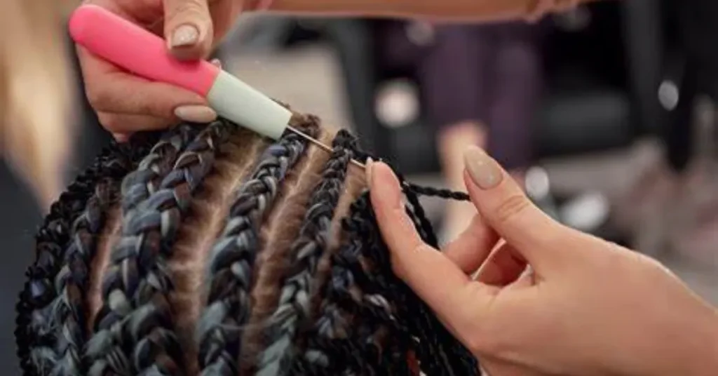Close-up of hands braiding stitch braids on a client's scalp using a crochet tool, showcasing detailed technique.